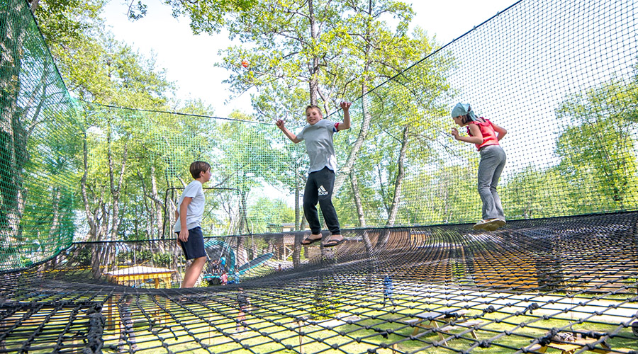 Cette image montre des enfants s'amusant sur un filet de trampoline dans un parc d'accrobranche, capturant l'énergie et la joie des activités en plein air entourées de nature verdoyante.