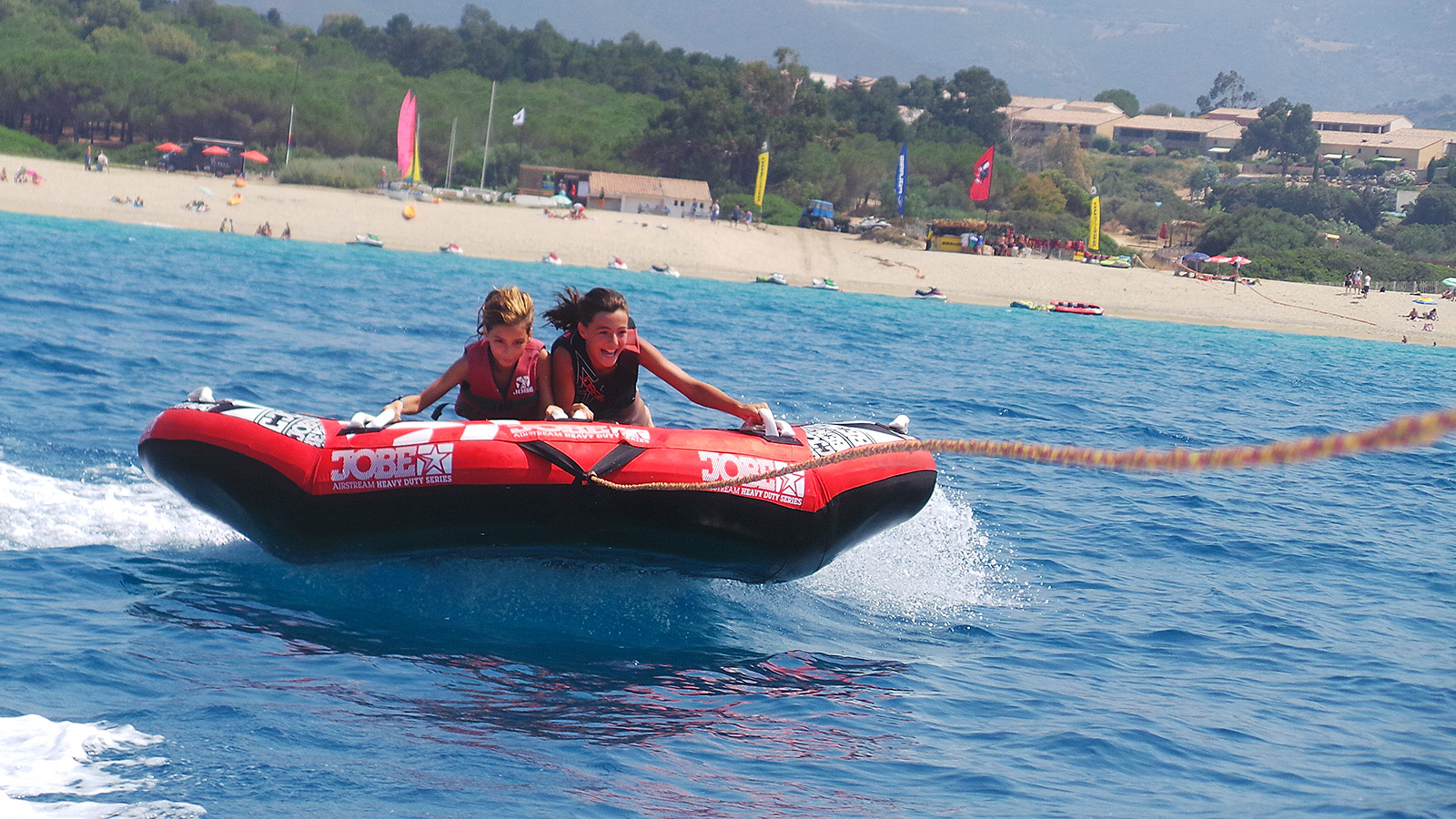 Enfants s'amusant sur un bateau-tube en mer Deux enfants rient de joie en mer, tirés par un bateau sur un tube lors de vacances d'été. Leurs rires résonnent avec les vagues sous le soleil brillant, capturant un moment magique d'insouciance sur la plage.
