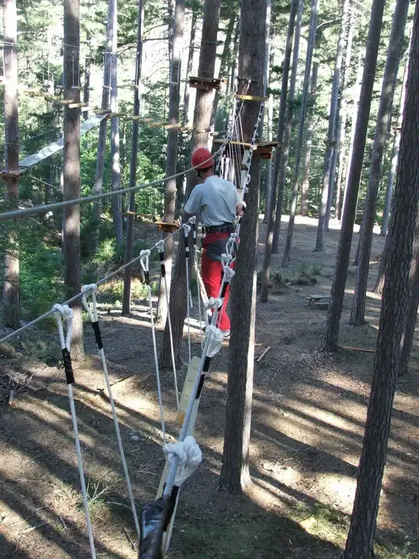 Un homme en casque rouge et pantalon rouge évolue sur un pont de singe en cordes dans une forêt de grands pins laricio en Corse, dans un parc de loisirs accrobranche.