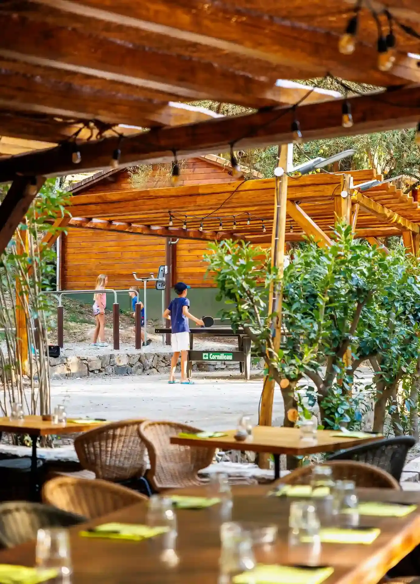 Enfants jouant au tennis de table sur une table Cornilleau sous une pergola en bois au camping Alivi di a Restonica à Corte.