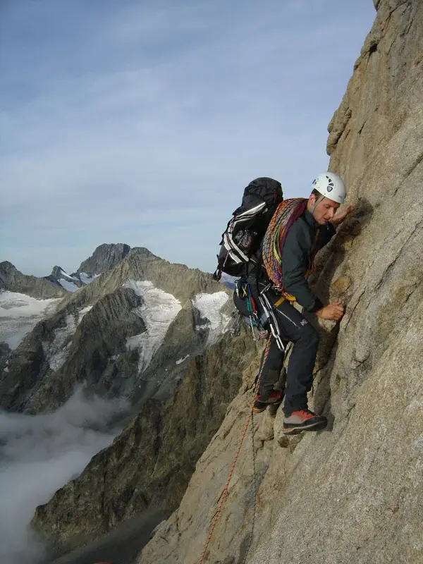 Un grimpeur équipé d'un casque blanc, d'un sac à dos et de cordes progresse sur une paroi de granit verticale et technique en Corse, avec un panorama de sommets enneigés et de nuages en arrière-plan.