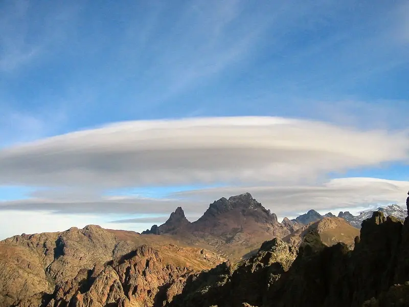 Un grand nuage lenticulaire blanc et plat plane au-dessus des crêtes montagneuses rocheuses et déchiquetées de la Corse, sous un ciel bleu avec des cirrus légers.