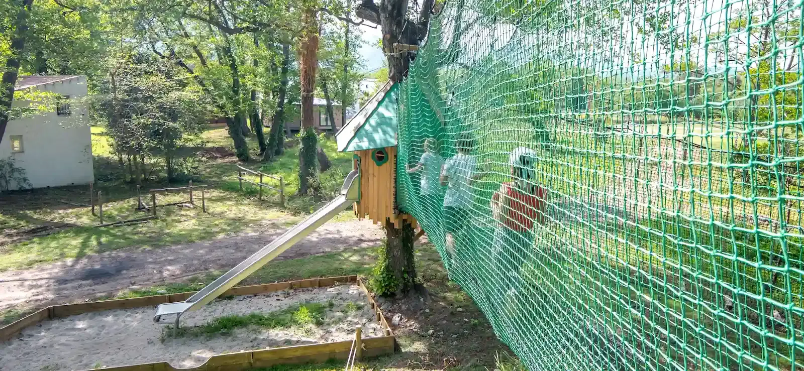 Vue d'un parcours accrobranche pour enfants au Camping Merendella, avec des filets de sécurité verts menant à une cabane dans les arbres et un toboggan en inox.