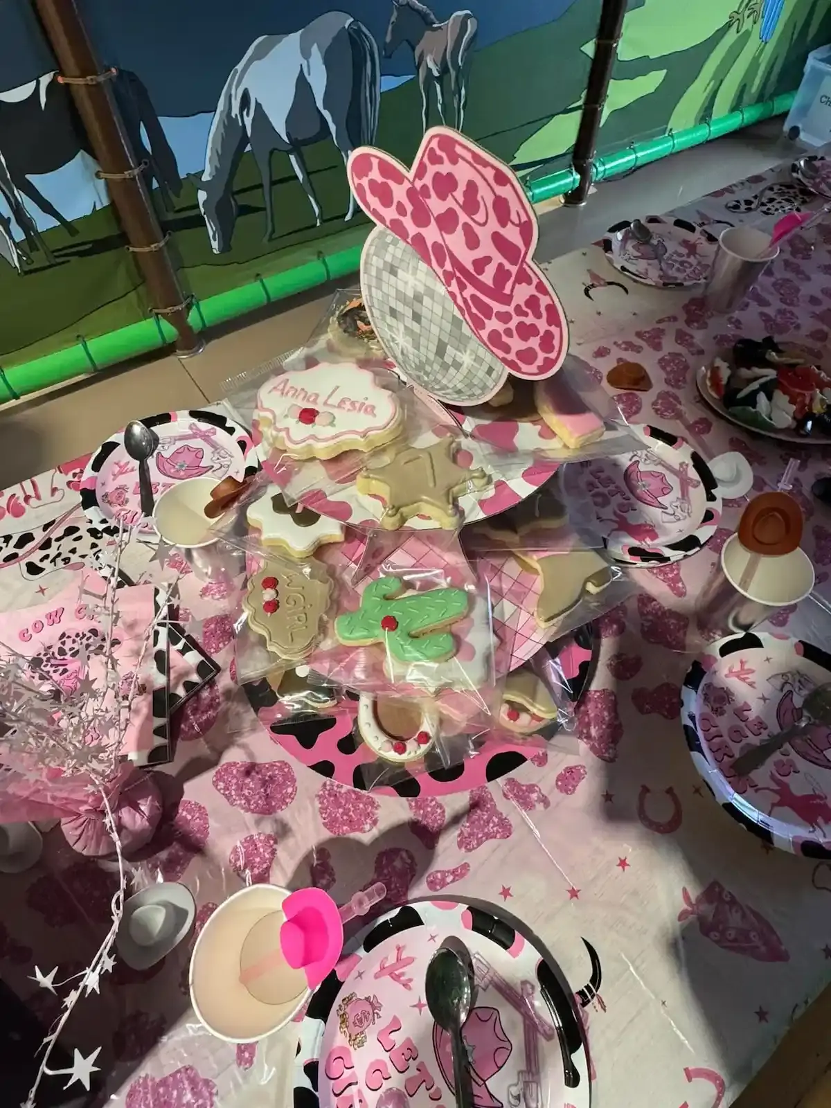 Table d'anniversaire décorée sur le thème Cow-girl avec nappe rose à motifs vache, biscuits personnalisés et chapeau rose.