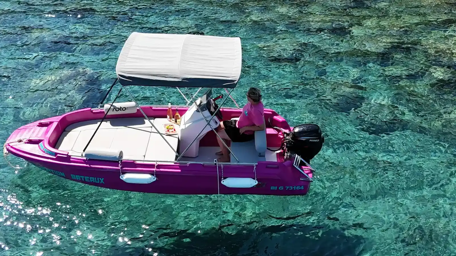 Bateau de location rose sans permis avec taud de soleil naviguant dans les eaux claires de la réserve de Scandola en Corse.