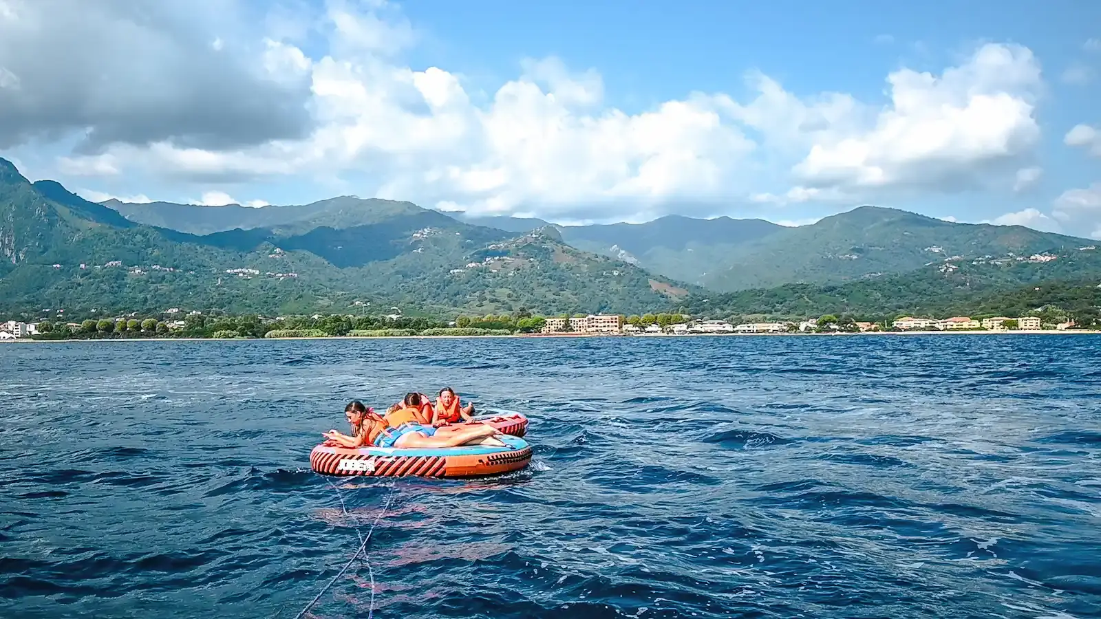 Groupe d'amis allongés sur une bouée plate ronde tractée par Costa Verde Aventure au large de Moriani-Plage, avec les montagnes de Haute-Corse en arrière-plan sous un ciel nuageux.