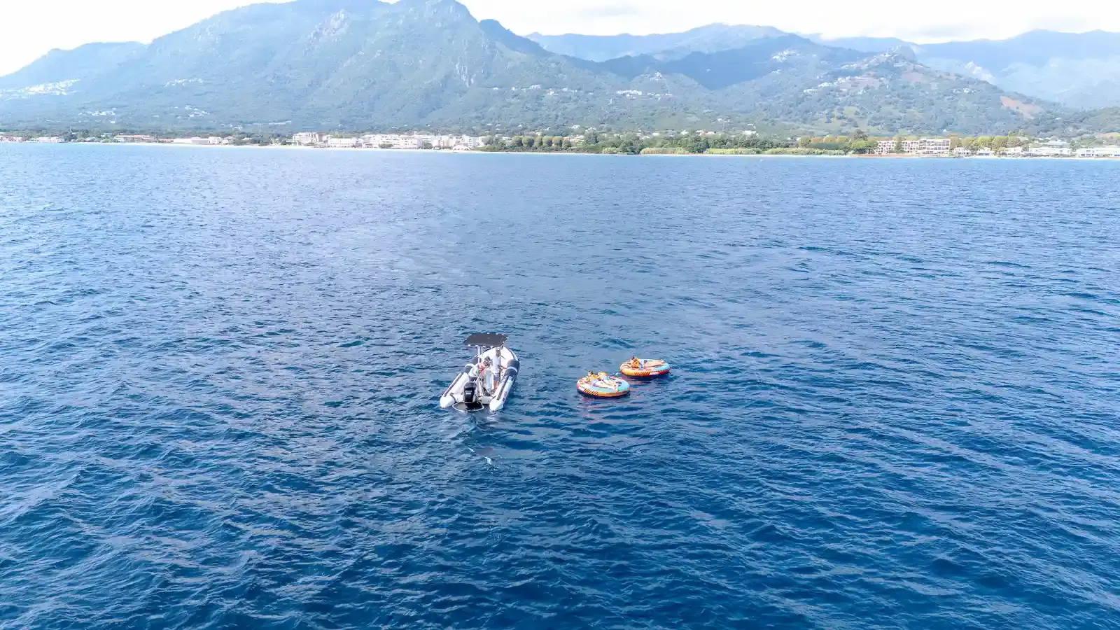 Vue aérienne par drone montrant un bateau à moteur de Costa Verde Aventure tractant deux bouées rondes avec des participants au large de la côte orientale de la Corse, près de Moriani.