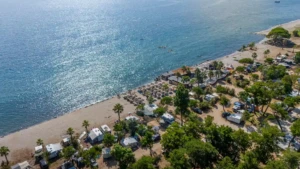 Vue aérienne du Camping Merendella avec emplacements ombragés, plage de sable fin et mer cristalline sous le soleil.