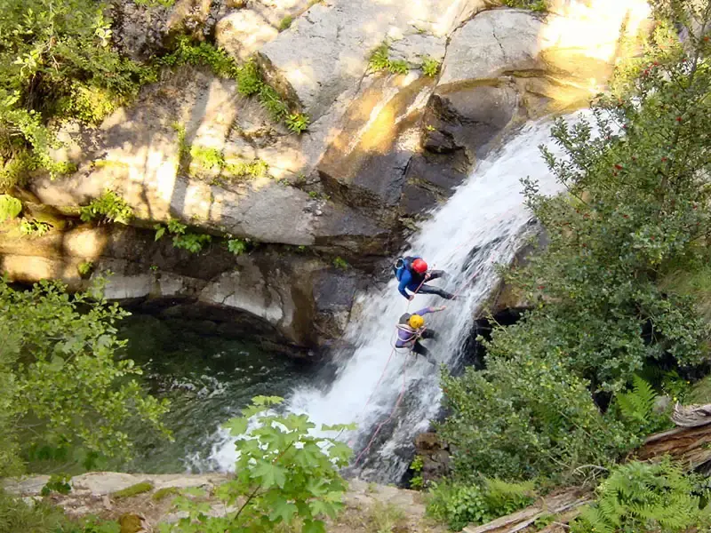 Deux personnes en équipement de canyoning (combinaison et casque) descendent en rappel le long d'une cascade d'eau vive se jetant dans une vasque verte, entourée de roches granitiques et de végétation.