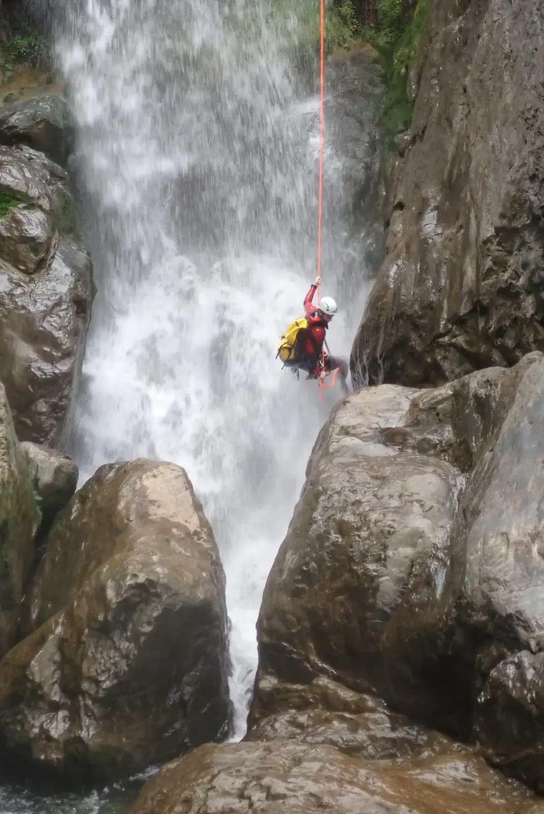 Un pratiquant de canyoning effectue une descente en rappel sur une corde orange devant une grande cascade d'eau vive en Haute-Corse.