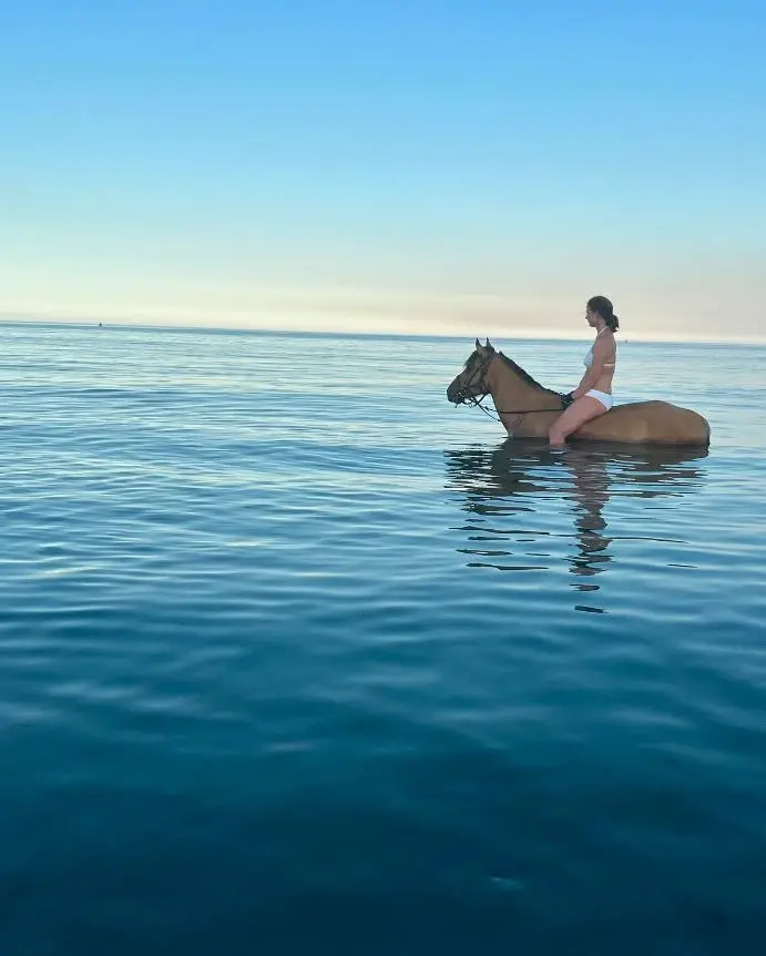 Femme en maillot de bain blanc à cheval au milieu d'une mer calme et bleue, loin du rivage.
