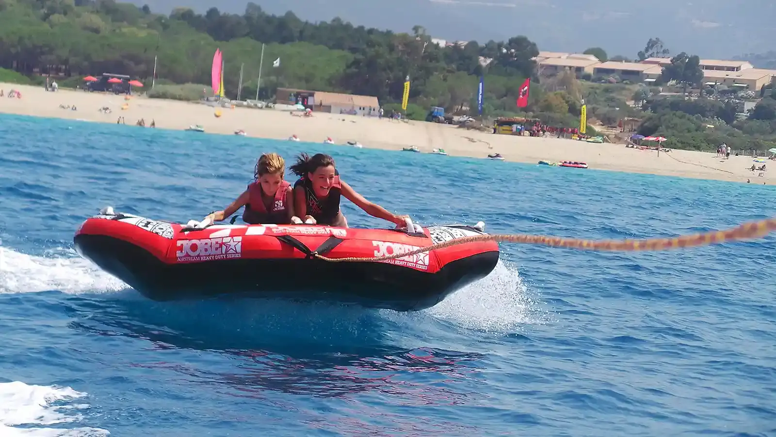Gros plan sur deux jeunes filles souriantes et s'agrippant à une bouée tractée Jobe Airstream rouge et noire, glissant sur l'eau turquoise près d'une plage animée de la Costa Verde.