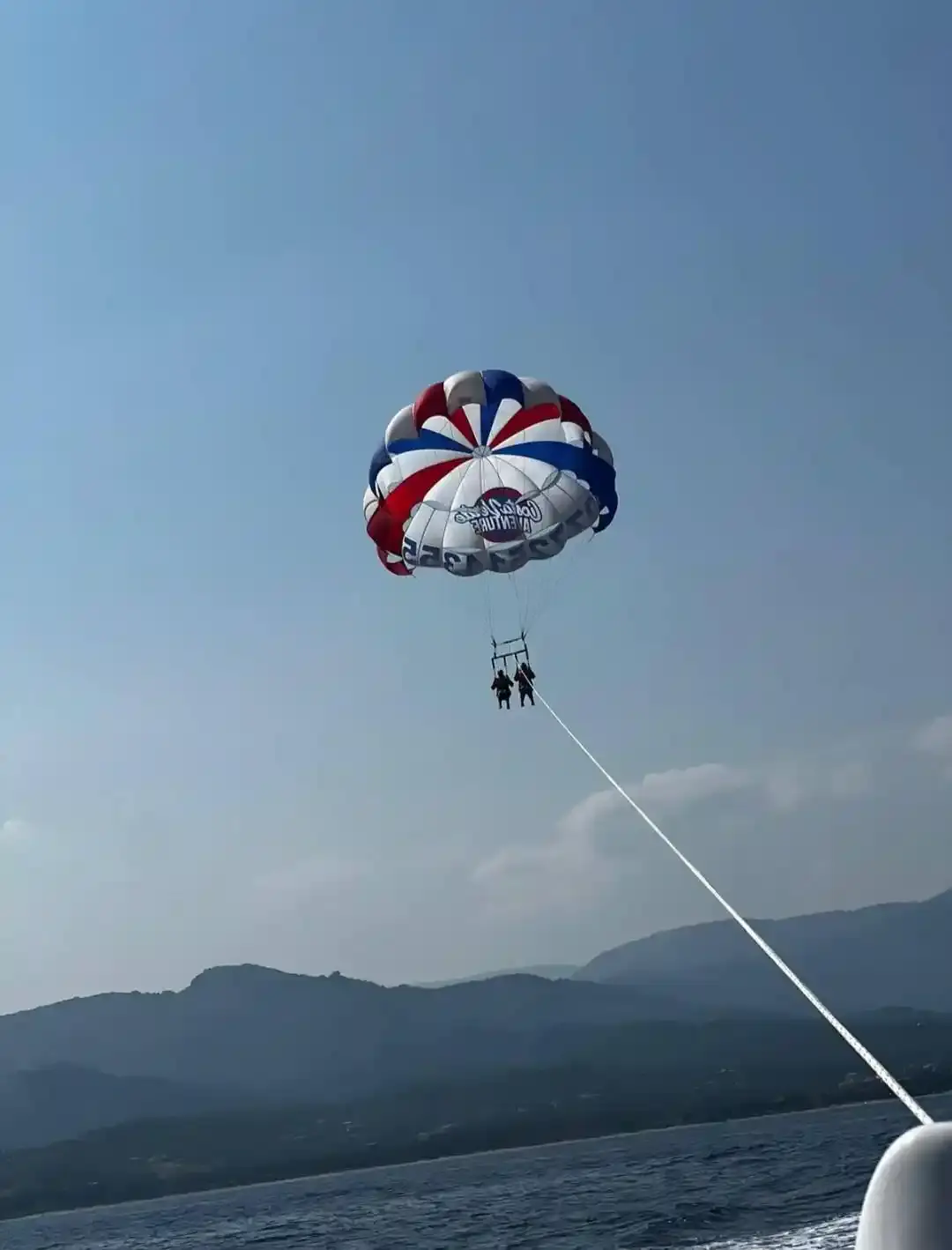 Vue en contre-plongée d'un parachute ascensionnel aux couleurs bleu, blanc, rouge, avec deux personnes suspendues, volant haut dans le ciel au-dessus de la Costa Verde, avec les montagnes de Corse à l'horizon.