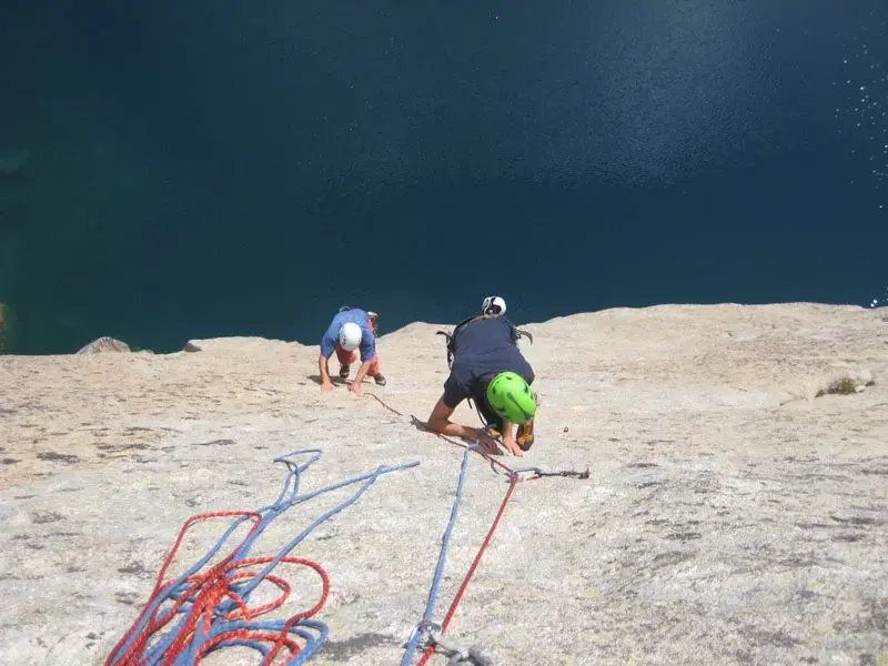 Vue du dessus de deux grimpeurs en casque (bleu et vert) progressant sur une paroi rocheuse de granit, directement au-dessus des eaux bleu émeraude d'un lac de montagne en Corse.