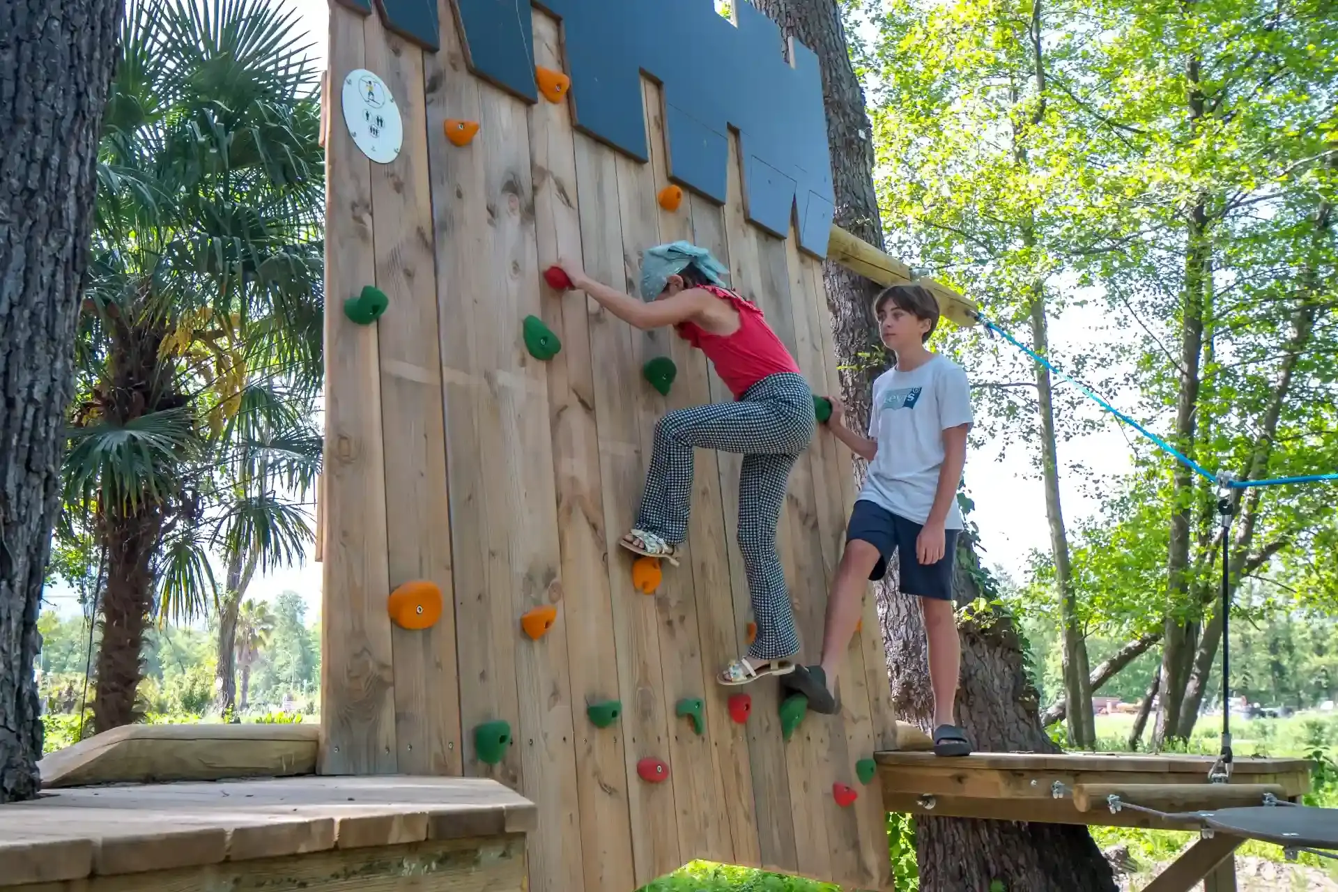 Une fille escalade un mur de grimpe en bois avec des prises colorées sous le regard encourageant d'un garçon, au Camping Merendella en Corse.