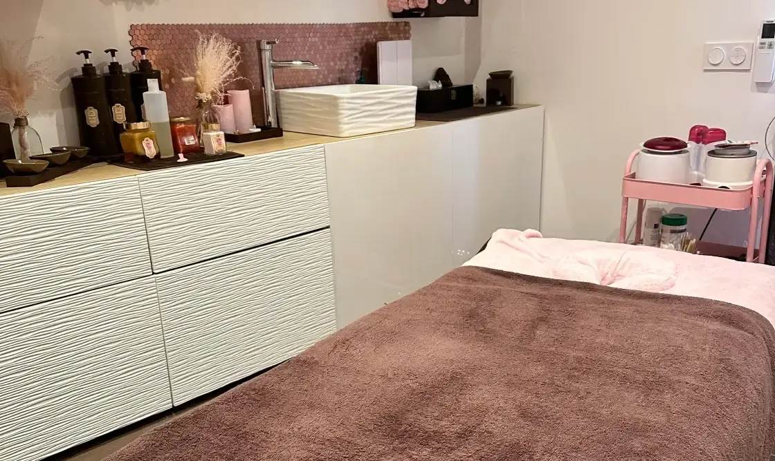 Intérieur d'une cabine de soin lumineuse avec table de massage préparée, rangements modernes et produits de beauté.