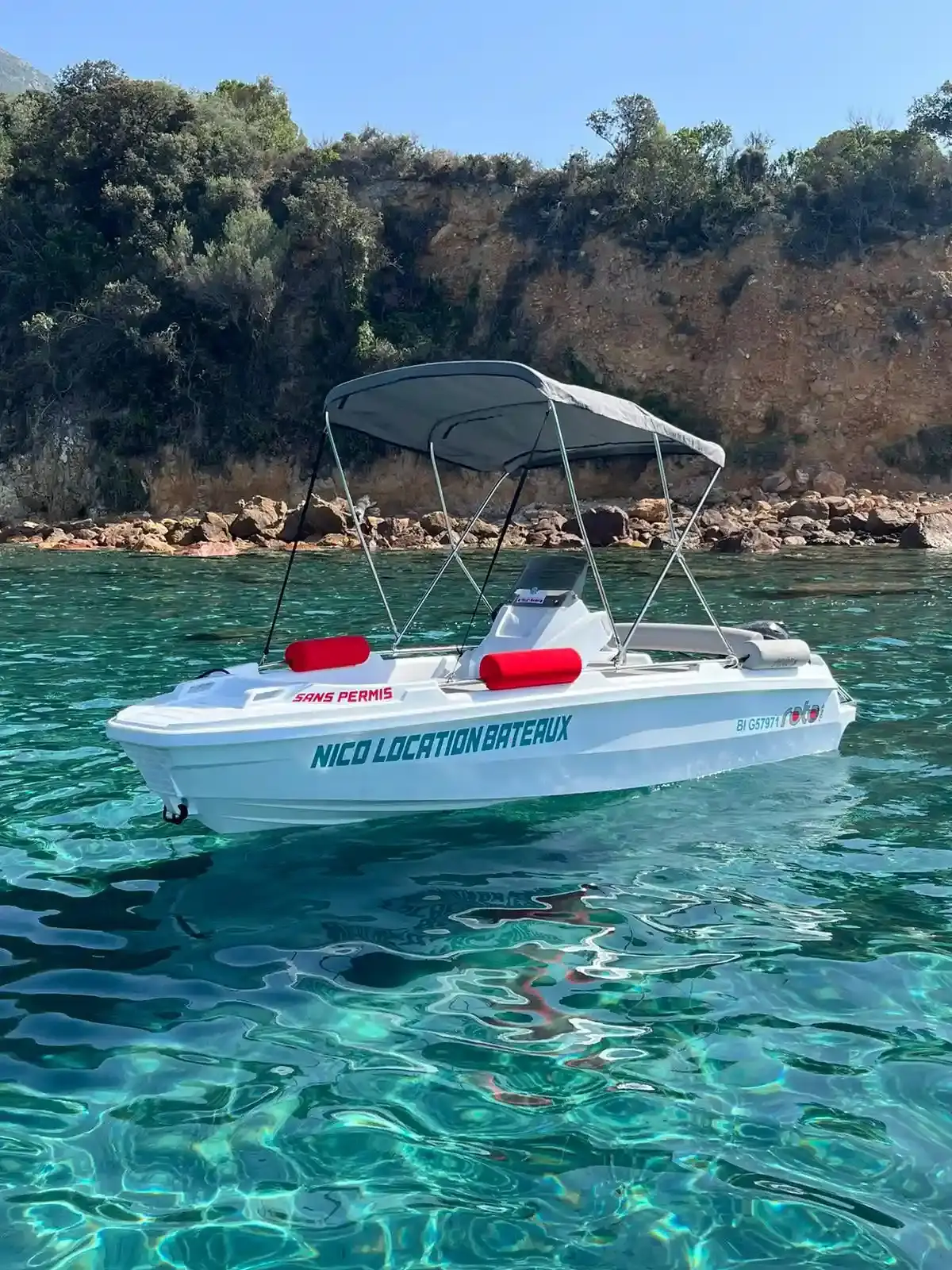 Bateau blanc sans permis Nico Location ancré sur une eau turquoise transparente avec une côte rocheuse en arrière-plan.
