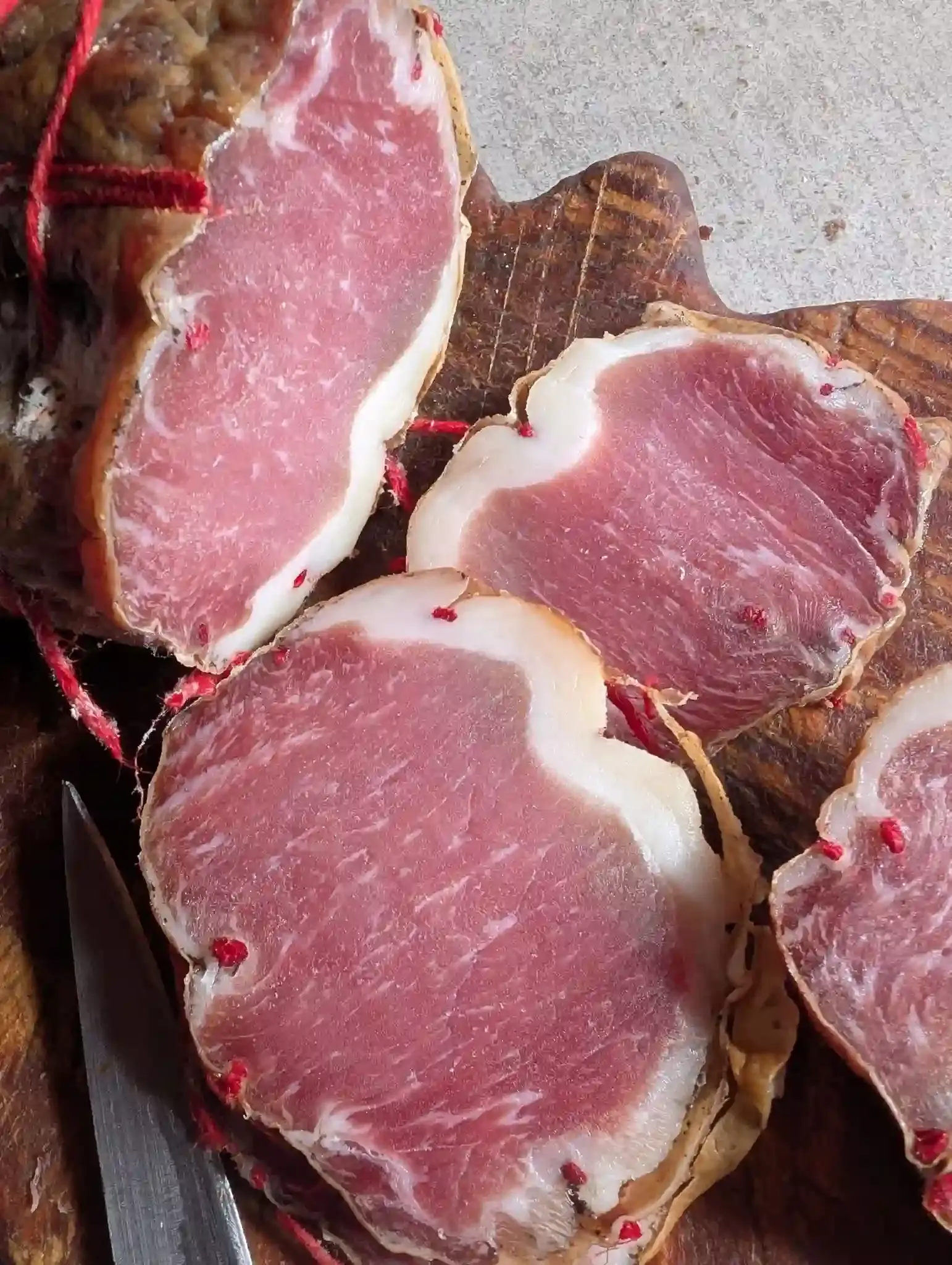 Des morceaux de lonzu corse artisanal, ficelés et prêts à être tranchés sur une planche de bois, avec un couteau à la charcuterie Gozzi.