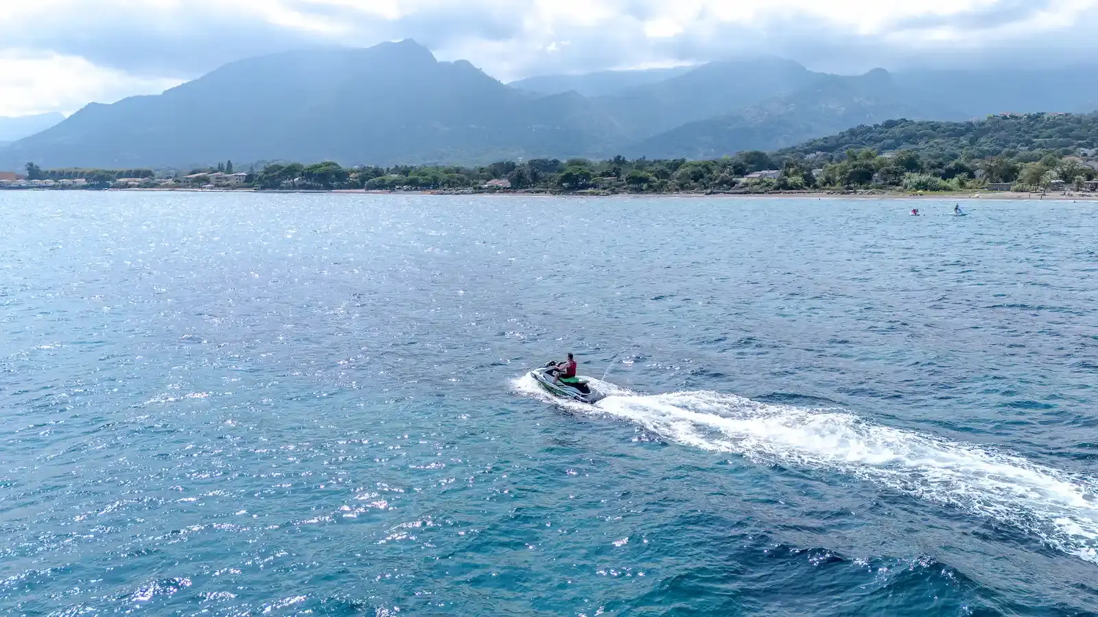 Panorama côtier de la Corse avec un jet ski s'éloignant vers les montagnes à l'horizon.
