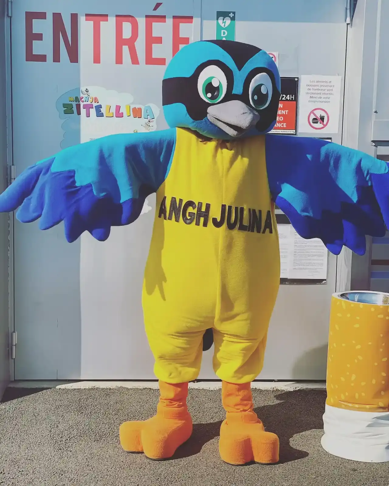 Personnage en costume de mascotte oiseau bleu et jaune nommé Anghjulina devant l'entrée du parc.