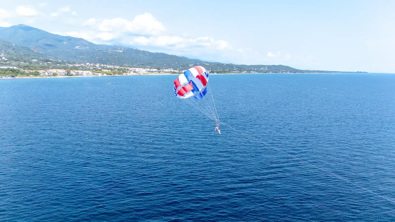 Vue aérienne par drone d'un vol en parachute ascensionnel tricolore au-dessus de la mer Tyrrhénienne, avec la côte de la Costa Verde et les montagnes de Haute-Corse en arrière-plan.