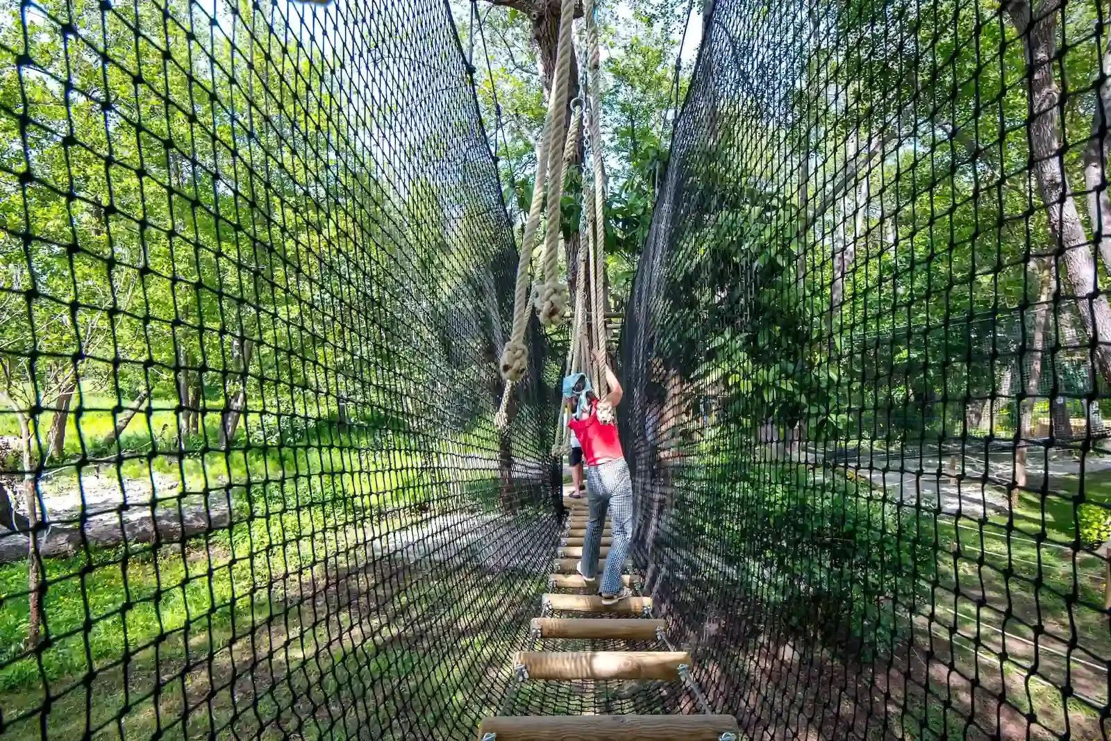 Vue en perspective d'un pont suspendu en tronçons de bois, entièrement sécurisé par de hauts filets de protection noirs au Camping Merendella.