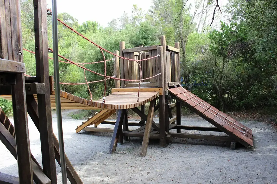 Aire de jeux pour enfants avec parcours d'équilibre en bois et pont de singe sous les arbres du camping.