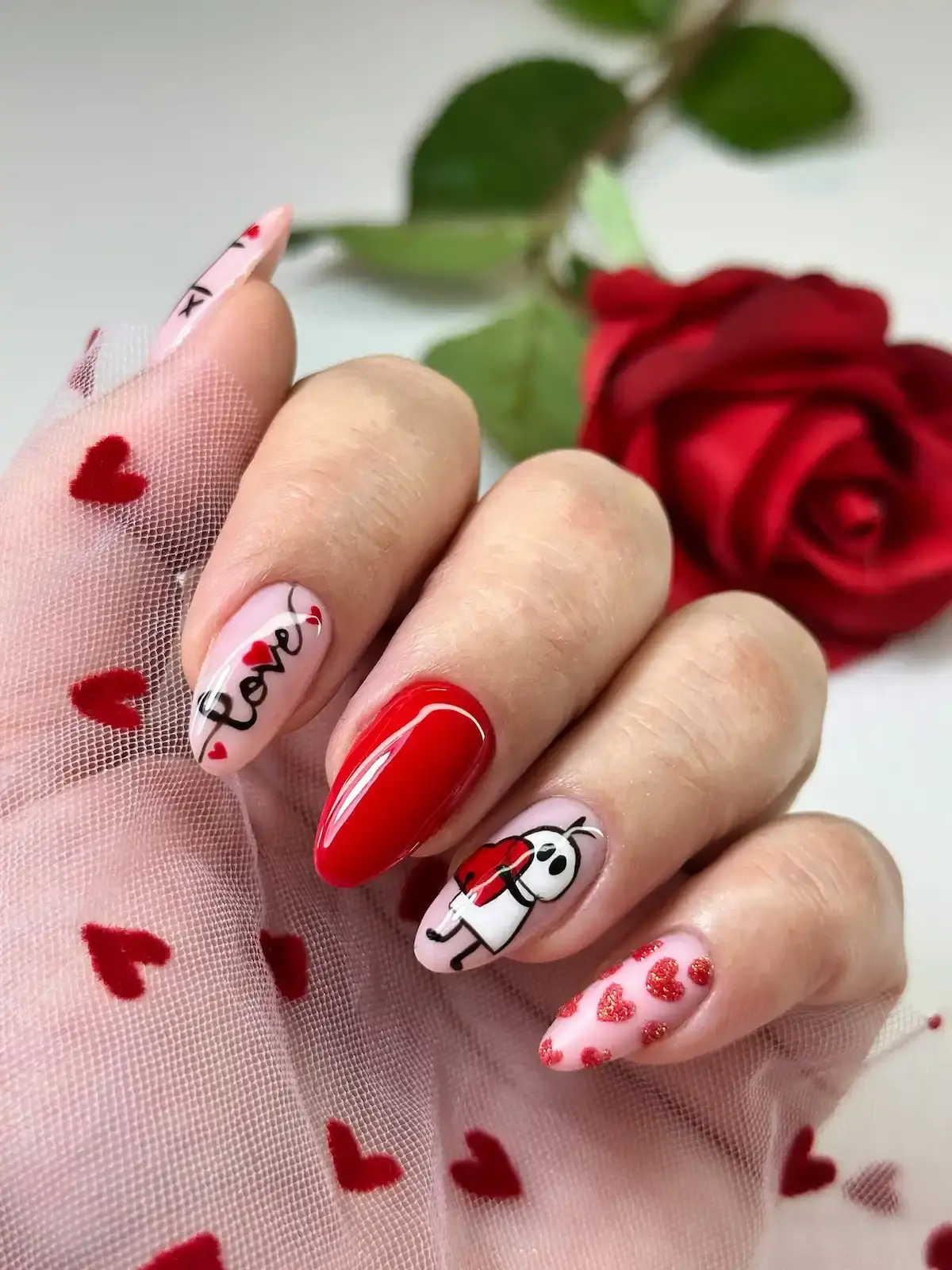 Nail art thématique avec vernis rouge brillant, motifs coeurs et personnage "Love" sur fond rose pâle