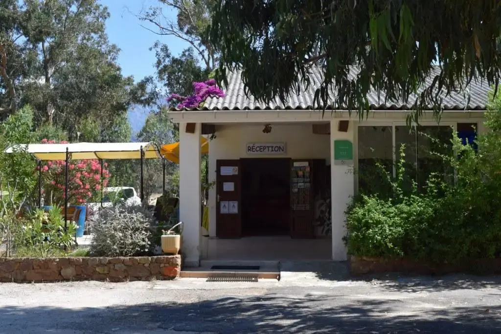 Bâtiment de la réception du Camping Paradella à Calvi, avec l'enseigne "RÉCEPTION", une terrasse couverte et de la végétation méditerranéenne.