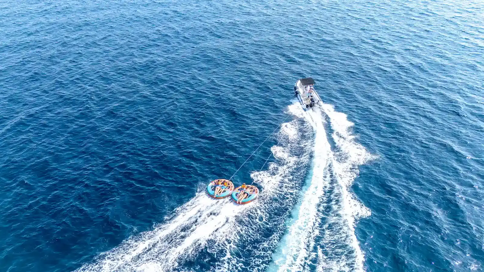 Vue aérienne en plongée d'un bateau semi-rigide de Costa Verde Aventure tractant deux bouées rondes orange avec des participants, créant un sillage blanc sur une mer bleu foncé, au large de la Costa Verde.