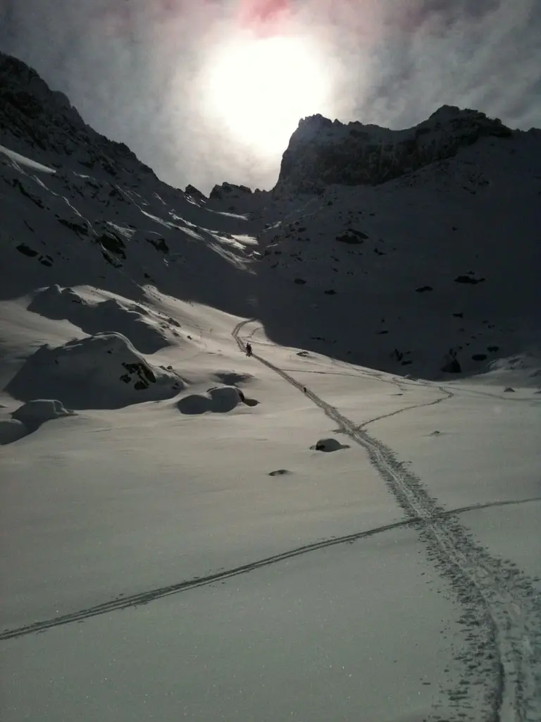 eux alpinistes remontant un couloir de neige escarpé à l'ombre de hautes parois rocheuses en Corse, sous un soleil hivernal éclatant créant un fort contre-jour.