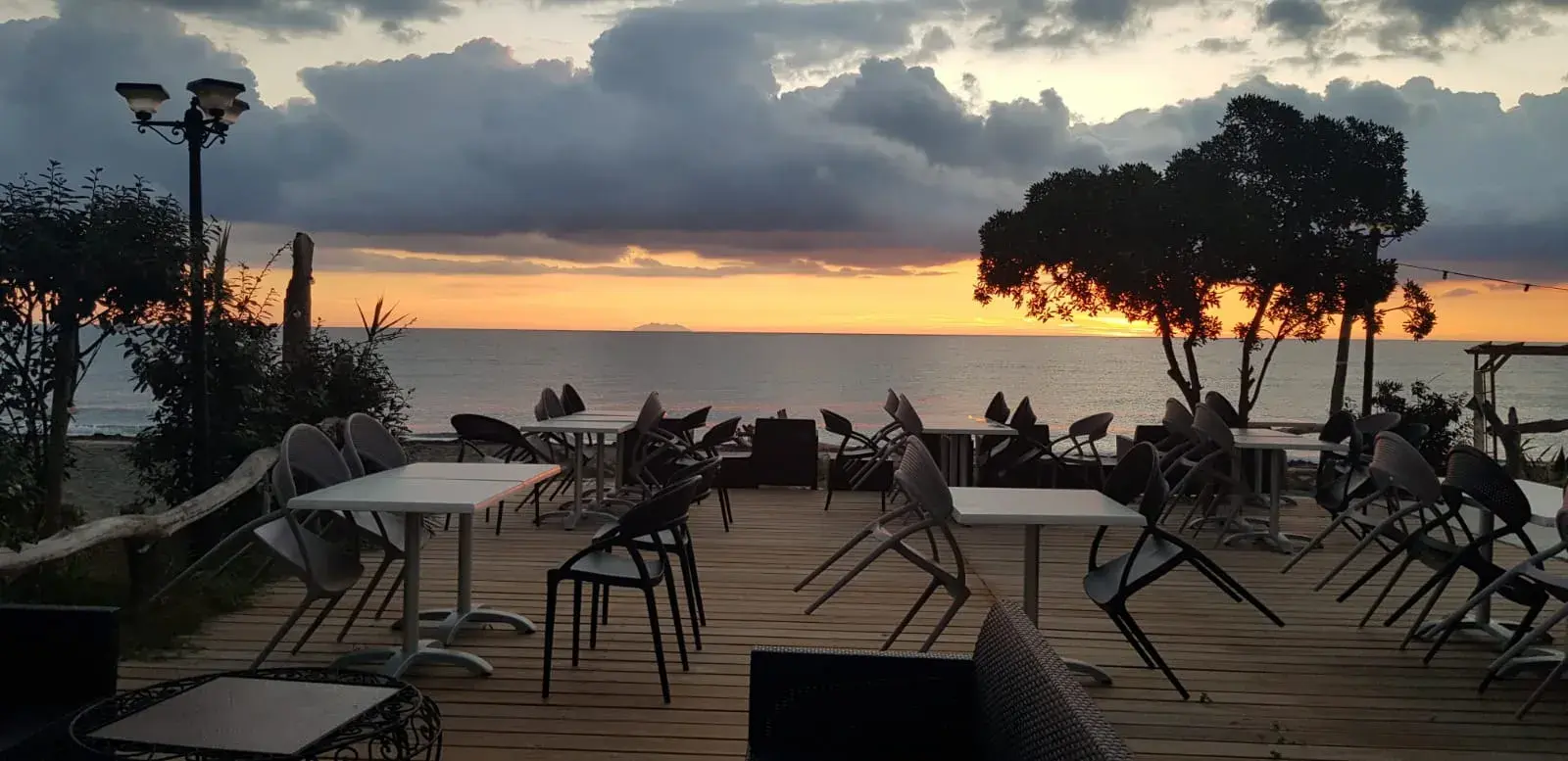 Tables de restaurant installées sur une terrasse en bois face à la mer sous un ciel de coucher de soleil nuageux.