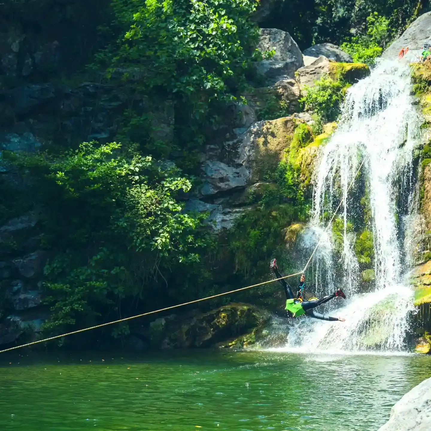 Une personne en tyrolienne survole une vasque d'eau verte et finit sa course près d'une cascade ensoleillée en Haute-Corse.