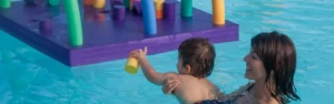 Jeune enfant soutenu par sa maman dans l'eau d'une piscine jouant avec des structures en mousse colorées.