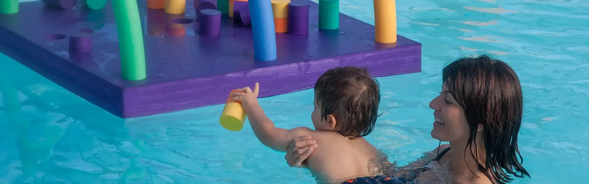 Jeune enfant soutenu par sa maman dans l'eau d'une piscine jouant avec des structures en mousse colorées.
