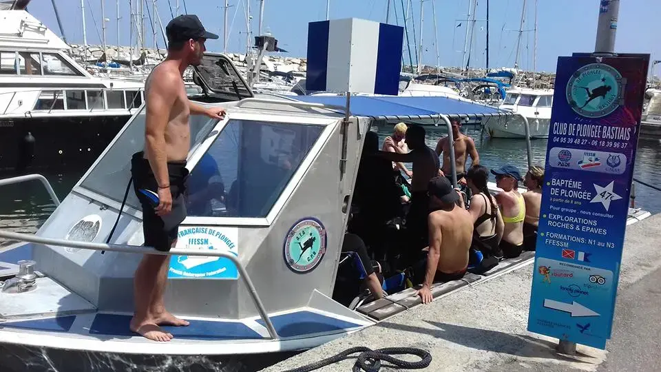 Groupe de plongeurs et moniteurs sur un bateau en aluminium devant le panneau des tarifs du Club de Plongée Bastiais sur le quai.
