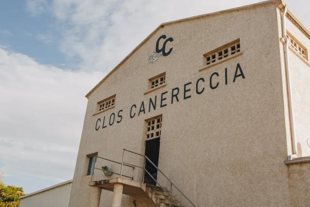 Façade beige du bâtiment principal du domaine viticole Clos Canereccia avec son logo CC noir sous un ciel bleu parsemé de nuages.