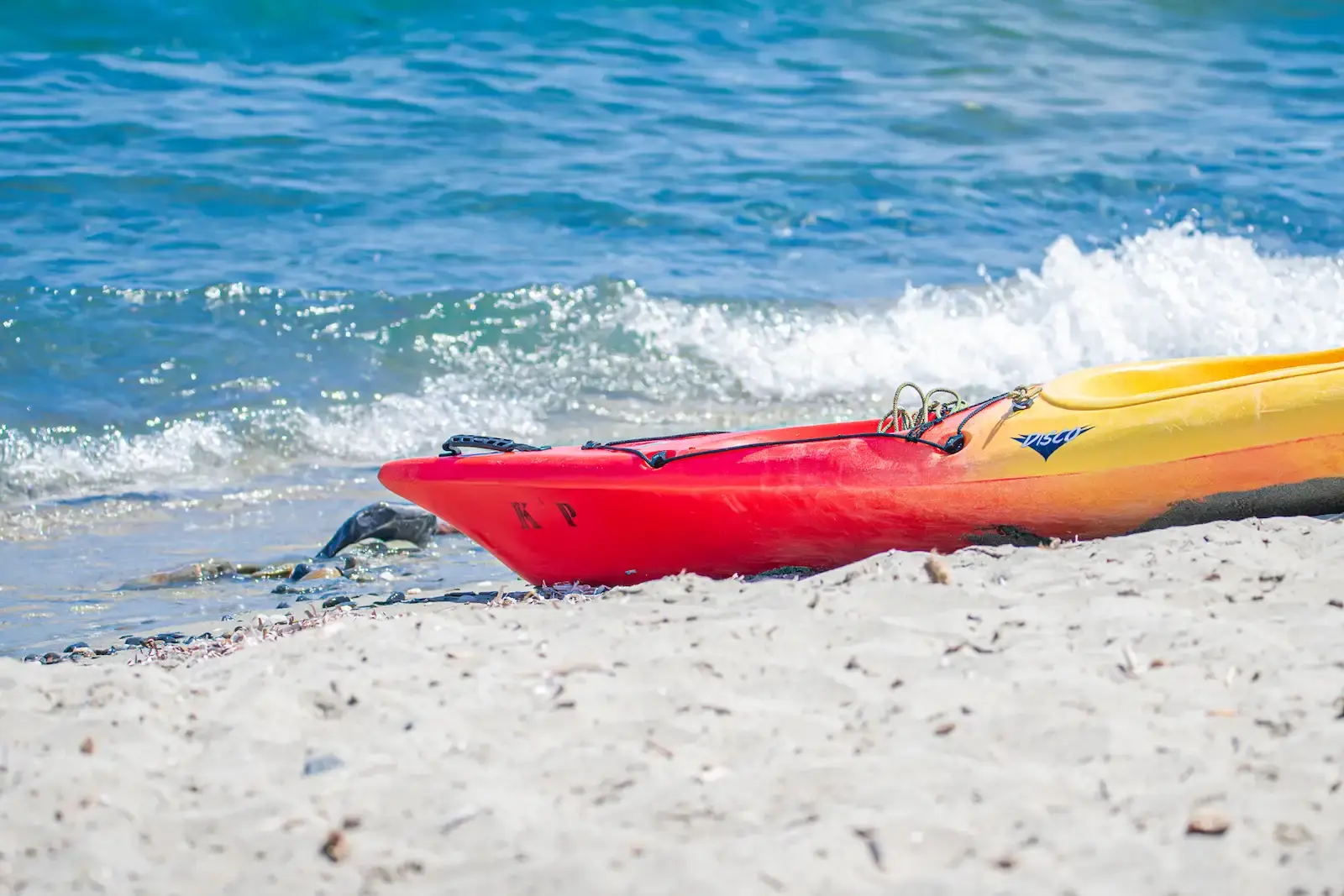 Kayak de mer rouge et jaune posé sur le sable blanc d'une plage en Corse avec de légères vagues en arrière-plan.
