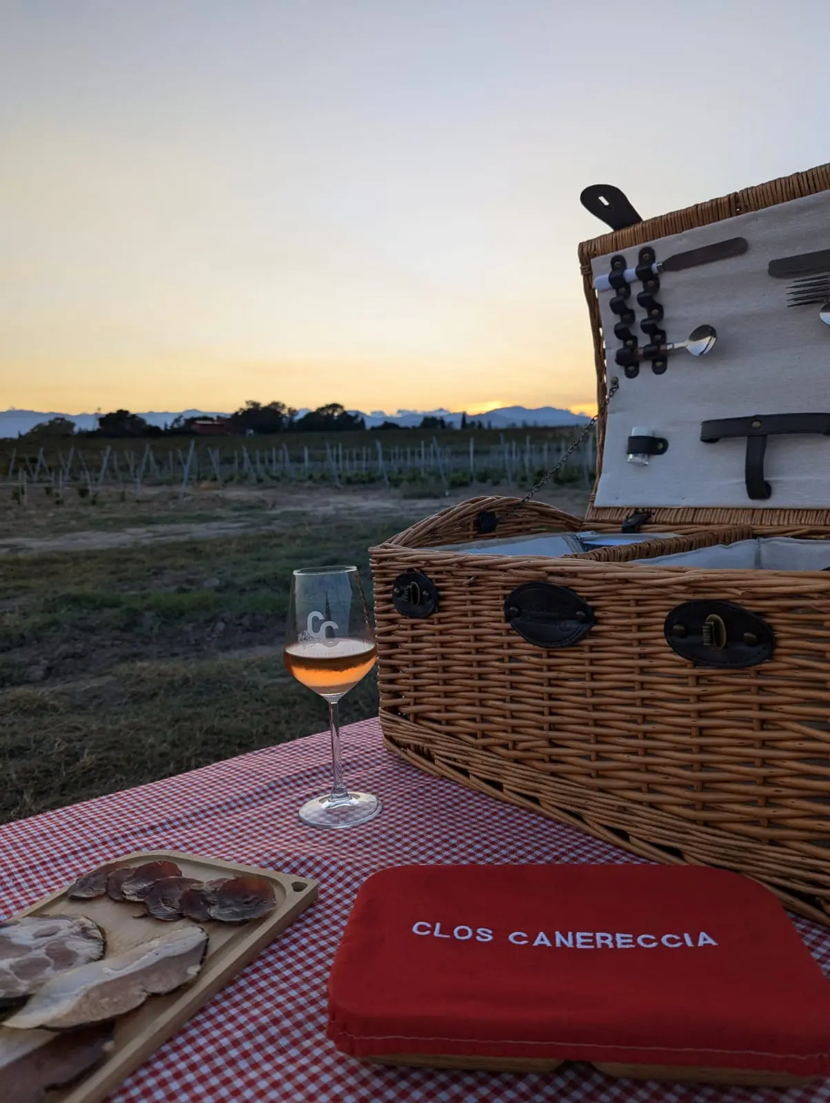 Panier de pique-nique en osier ouvert sur une nappe à carreaux rouges avec un verre de vin rosé face au coucher de soleil sur les vignes.