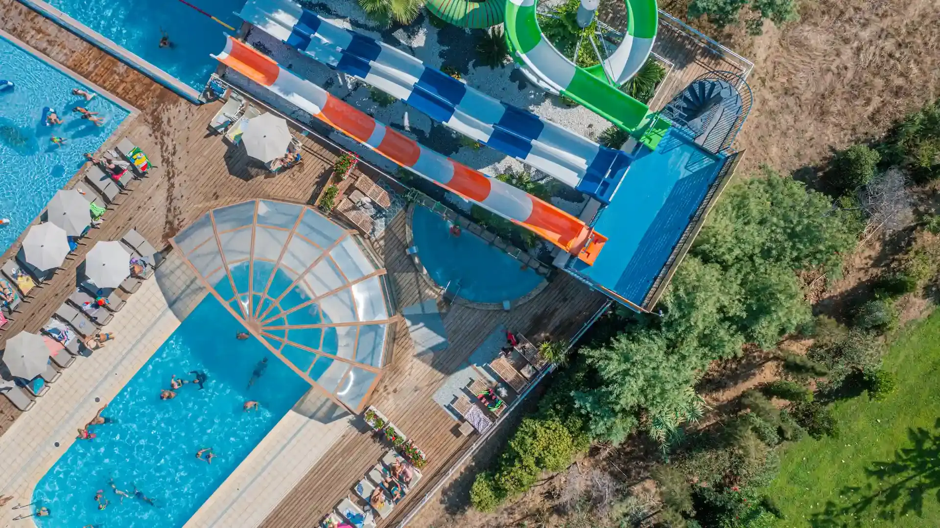 Vue aérienne d'un parc aquatique haut de gamme en Haute-Corse avec toboggans géants colorés, piscine chauffée avec dôme vitré et vacanciers sur des transats.