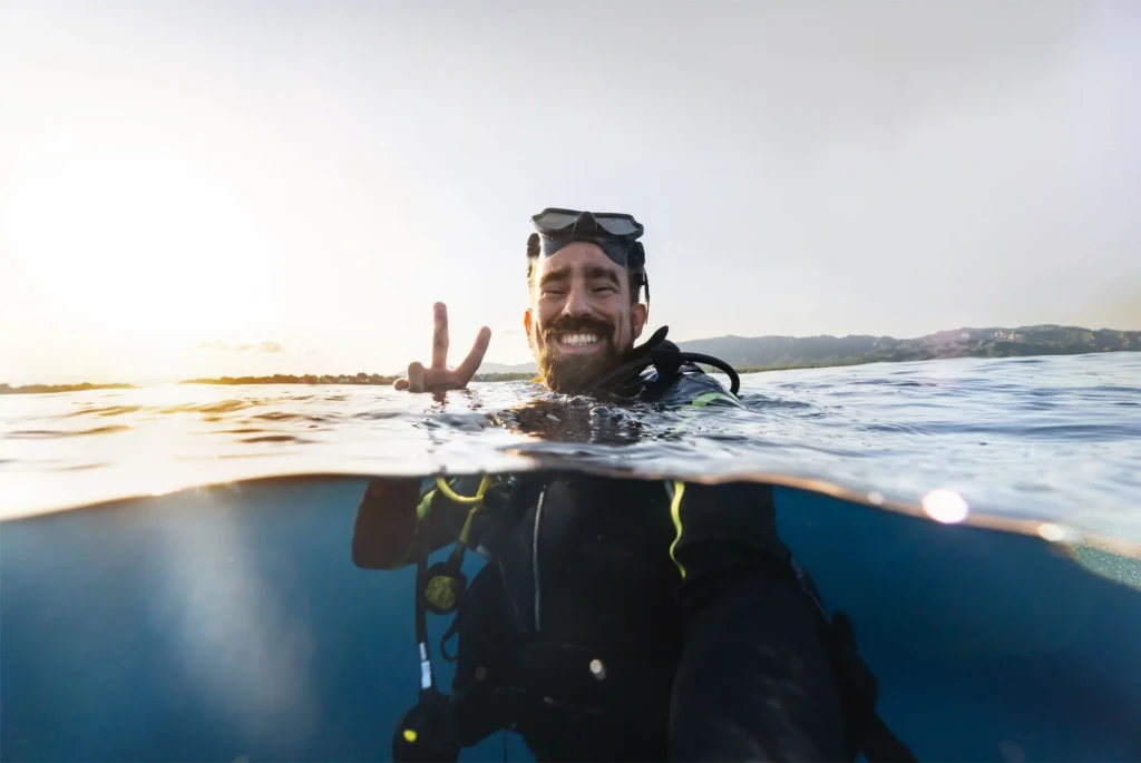 Plongeur souriant équipé d'un masque et d'un détendeur faisant le signe de la paix à la surface de l'eau turquoise en Haute-Corse.