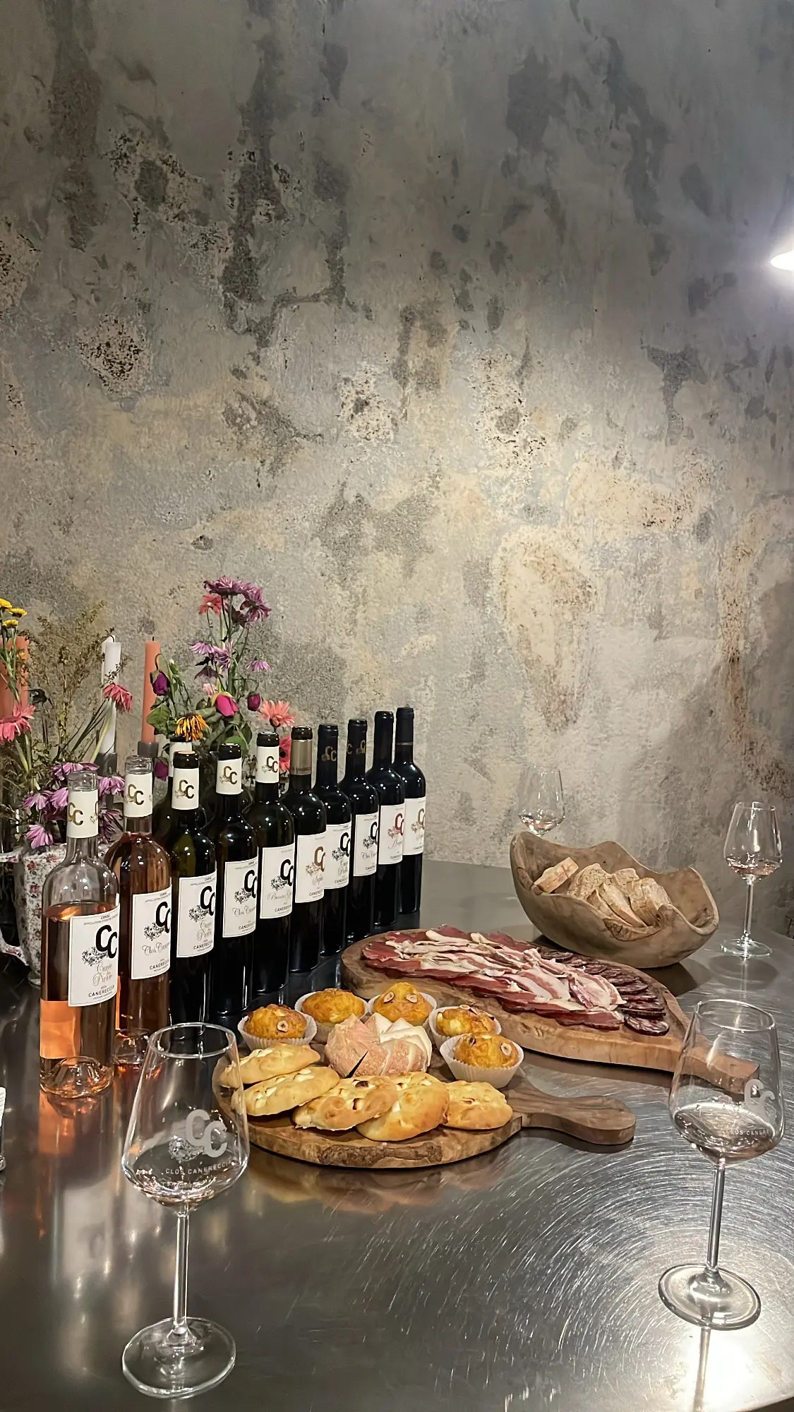 Alignement de bouteilles de vin blanc, rosé et rouge Clos Canereccia sur un comptoir en inox avec verres de dégustation et planches de charcuterie.