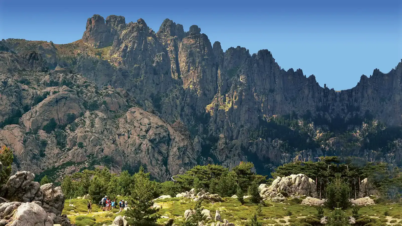 Groupe de randonneurs avec sacs à dos progressant sur un sentier verdoyant au pied des sommets rocheux des Aiguilles de Bavella en Corse.