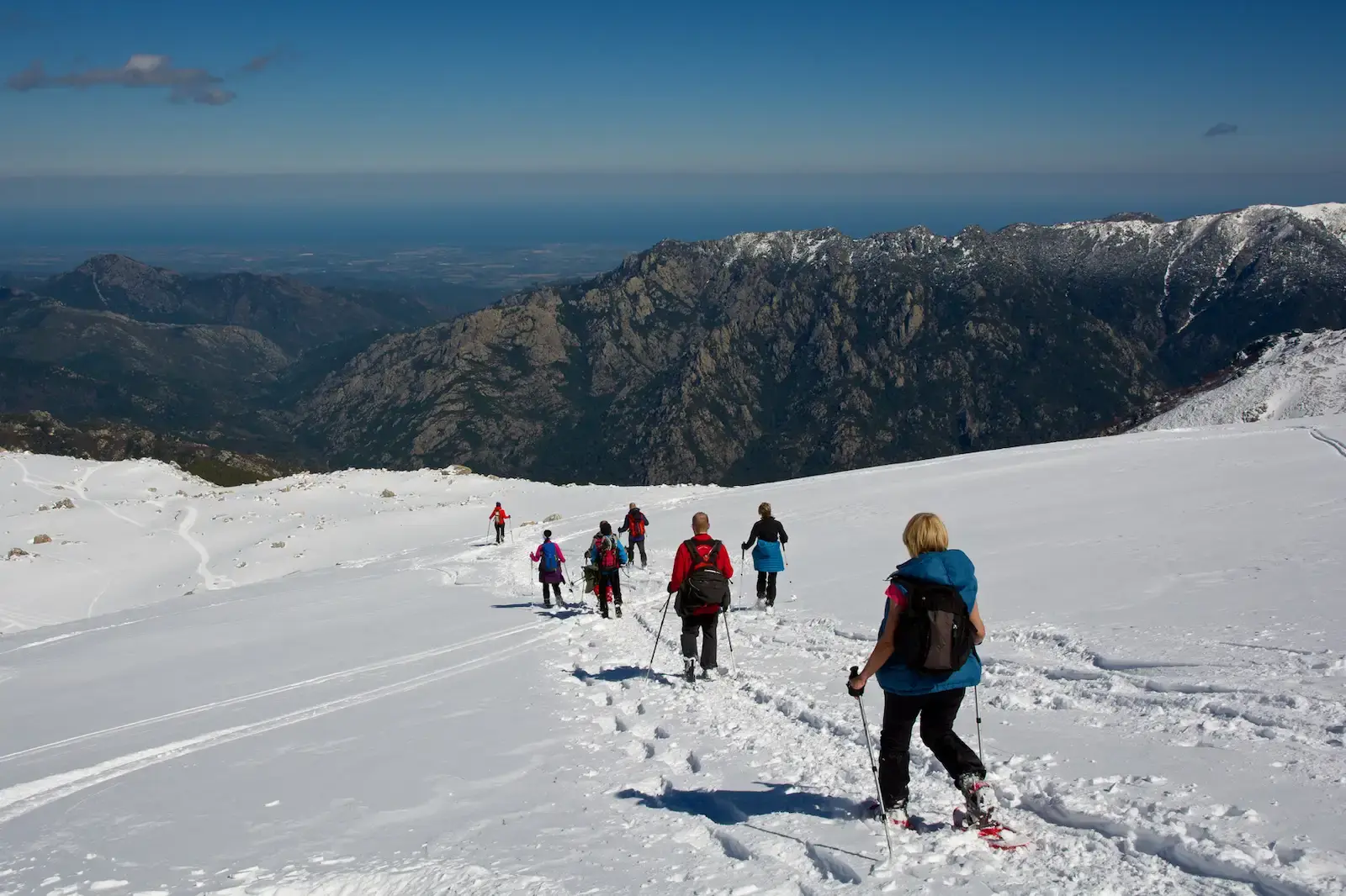 Groupe de personnes pratiquant la randonnée en raquettes sur une pente enneigée avec vue sur les montagnes au loin.