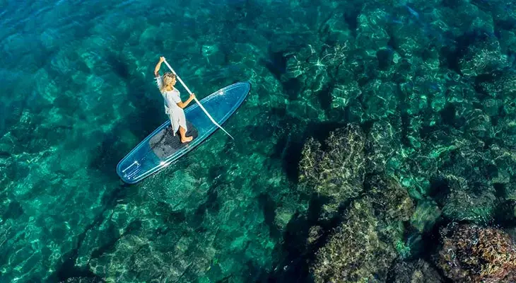 Femme pratiquant le stand up paddle transparent sur une eau turquoise cristalline permettant de voir les rochers sous-marins sur la côte corse.