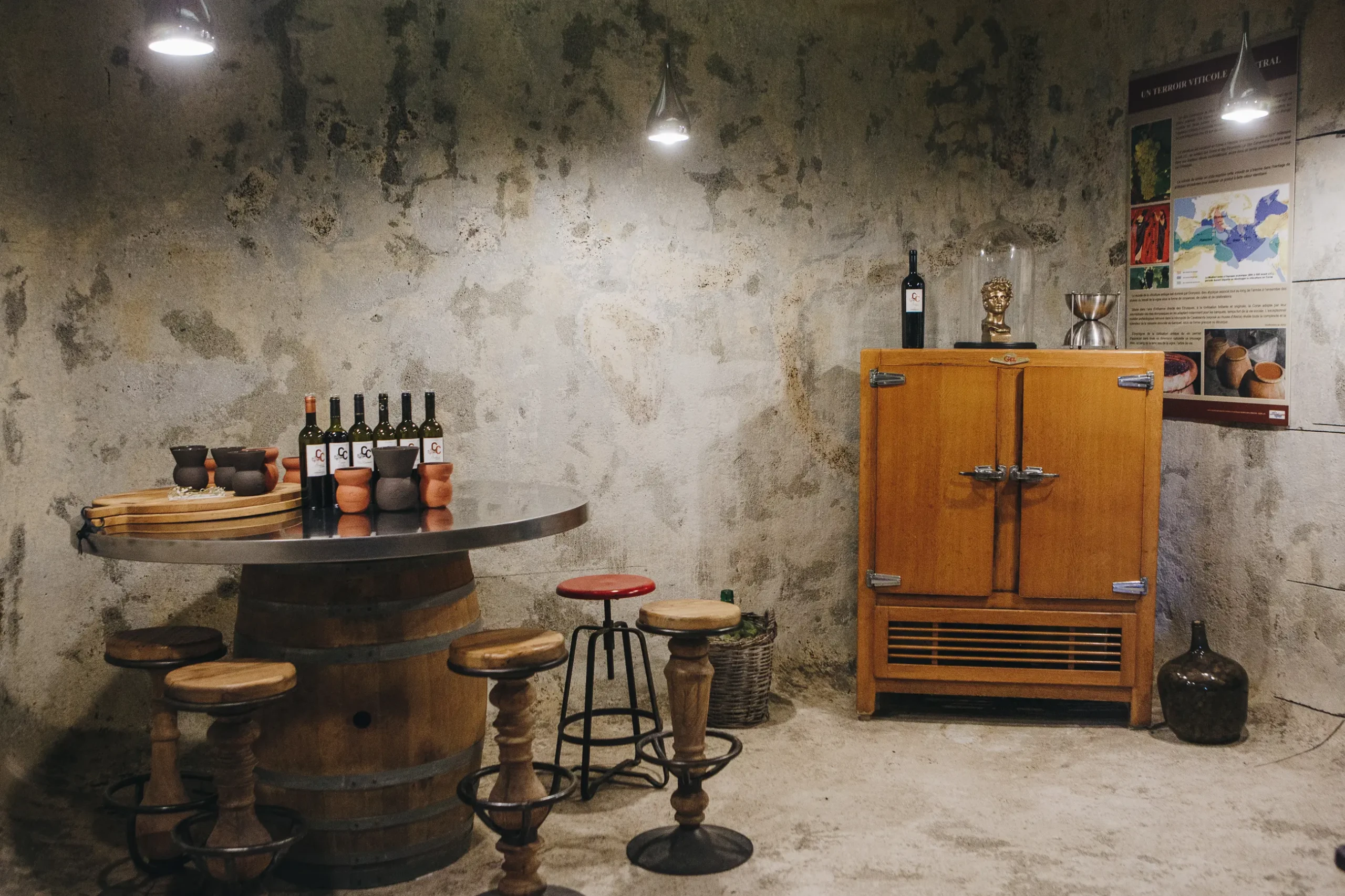 Espace de dégustation aménagé dans une cave en pierre avec une table tonneau, des tabourets en bois et des bouteilles de vin du Clos Canereccia.